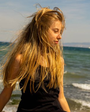 Siyah bir yaz elbisesi giymiş, kumsal arka planında rüzgarda savrulan sarışın bir kızın yakın plan portresi.