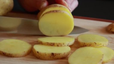 Kadının elleri tahta kesme tahtasının üzerinde çiğ patatesleri ince daireler halinde dilimliyor.