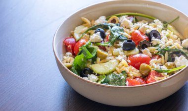  Taze sebzelerden, siyah zeytinlerden ve peynirden yapılmış bir kasede servis edilen sağlıklı orzo salatası.