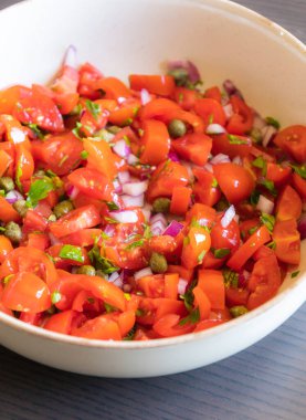 Çeri domatesli, kırmızı soğanlı, kapari ve maydanozlu taze salsa sosu bir kasede servis edilir. Malzemelerin renkli karışımı canlı ve sağlıklı bir görünüm veriyor.