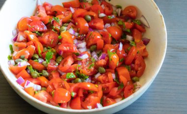 Çeri domatesli, kırmızı soğanlı, kapari ve maydanozlu taze salsa sosu bir kasede servis edilir. Malzemelerin renkli karışımı canlı ve sağlıklı bir görünüm veriyor.