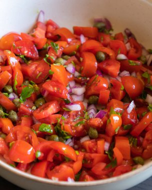 Çeri domatesli, kırmızı soğanlı, kapari ve maydanozlu taze salsa sosu bir kasede servis edilir. Malzemelerin renkli karışımı canlı ve sağlıklı bir görünüm veriyor.