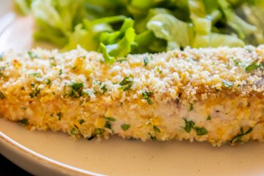 Fırında pişirilmiş somon filetosunun yakın çekimi. Bir tabakta sade yeşil salata ile servis ediliyor. Somon, altın ve çıtır çıtır bir kabuğa sahiptir ve taze yeşilliklerle eşleştirilir.