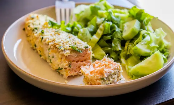 Fırında pişirilmiş somon filetosunun yakın çekimi. Bir tabakta sade yeşil salata ile servis ediliyor. Somon, altın ve çıtır çıtır bir kabuğa sahiptir ve taze yeşilliklerle eşleştirilir.