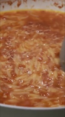 Tavada makarna pişirirken çekilmiş dikey yakın çekim videosu. Küçük makarna taneleri sıcak sıvıda kaynayarak sıcak ve iştah açıcı bir mutfak sahnesi oluşturuyor.