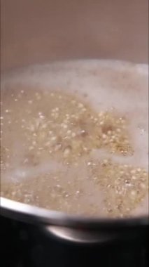 Sobanın üzerinde küçük bir tencerede kinoanın pişerken çekilmiş dikey yakın çekim videosu. Tahıllar suda yavaşça kaynayıp sağlıklı ve doğal gıda hazırlama sürecini sergiliyor.