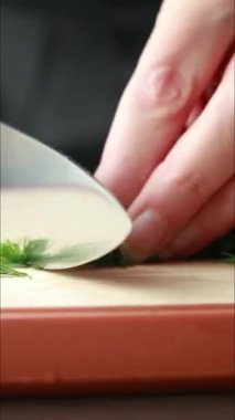 Tahta bir tahtada taze dereotu kesen bir bıçağı gösteren dikey yakın plan bir video. Yeşil bitki ince ince dilimlenmiş, hızlı ve temiz kesikler.