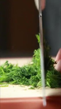 Tahta bir tahtada taze dereotu kesen bir bıçağı gösteren dikey yakın plan bir video. Yeşil bitki ince ince dilimlenmiş, hızlı ve temiz kesikler.