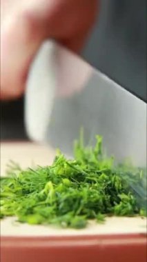 Tahta bir tahtada taze dereotu kesen bir bıçağı gösteren dikey yakın plan bir video. Yeşil bitki ince ince dilimlenmiş, hızlı ve temiz kesikler.