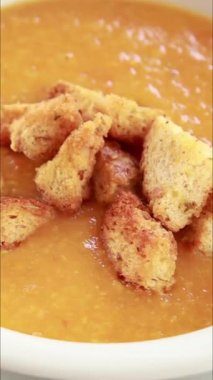 Bir kasede sıcak domates ve kabak çorbası servis eden ve üstüne ev yapımı kızarmış ekmek koyan ellerin yakın plan görüntülerini gösteren dikey bir video.