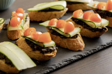 Yunan usulü üzerine zeytin ezmesi, salatalık dilimleri, vişne domatesleri ve peynir konmuş crostini resmi.