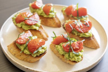 Kavrulmuş kiraz domatesli avokado crostini ve taze fesleğen. Isırık büyüklüğünde bir parti aperatifi olarak servis ediliyor. Yemek blogları ve eğlenceli fikirler için mükemmel.