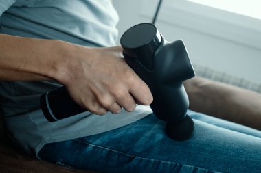Kot pantolon giyen beyaz bir adam, kahverengi bir koltukta otururken kalçasının kaslarına masaj yapmak için masaj tabancası kullanıyor.