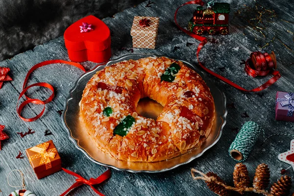 a roscon de reyes, the spanish king cake eaten traditionally on epiphany day, served on a silver plate, placed on a gray rustic table, next to some christmas ornaments, gifts and other seasonal items