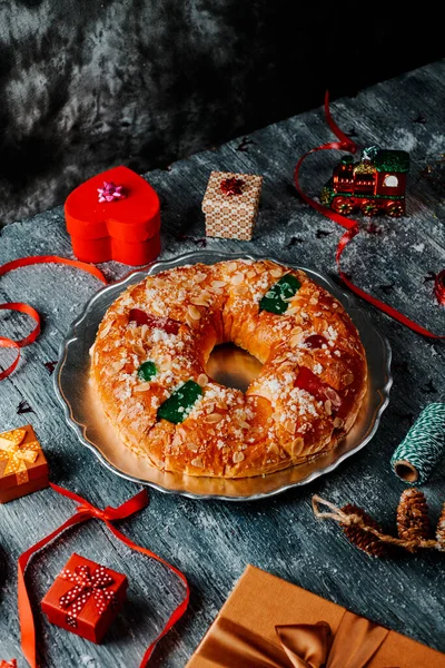 スペインの典型的な王のケーキ 伝統的にエピファニーの日に食べられるローソン レイエス いくつかのクリスマスの装飾品 贈り物 その他の季節のアイテムに囲まれた灰色の素朴なテーブルで — ストック写真