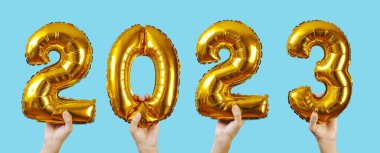 Yeni yıl olarak 2023 sayısını oluşturan bazı altın sayı şekilli balonları tutan eller mavi bir zemin üzerinde, web pankartı olarak kullanmak üzere panoramik bir formatta