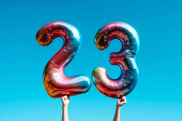 closeup of a man showing two number-shaped balloons forming the number 23, against the blue sky