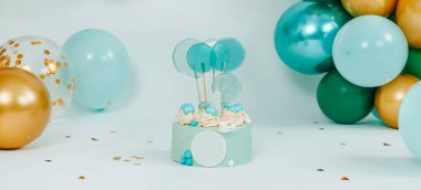 a blue cake on a white background in front of an elegant arrangement of balloons of different colors for a birthday party a wedding or a baby cake smash, in a panoramic format to use as web banner