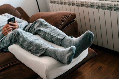 a man leans his legs on a leg elevation pillow, made of memory foam, while is resting on the sofa clipart