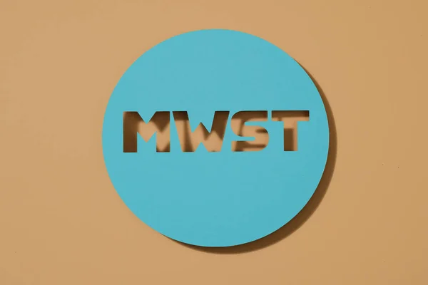 a blue paper sign with the german acronym MWST, for MehrwertSteuer, value-added tax, on a brown background