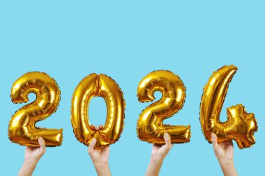 Yeni yıl olarak 2024 sayısını oluşturan altın şekilli balonları tutan eller mavi bir arka planda, üzerinde boş bir alan var.