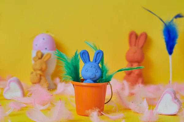 一只蓝色的复活节兔子 在橙色的水桶里 周围是粉色的羽毛和粉色的复活节蛋 还有其他一些黄色背景的复活节兔子 — 图库照片