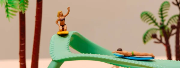 a miniature woman and a miniature man each on their surfing board, on a green flip-flop, against a pale brown background, in a panoramic format to use as web banner