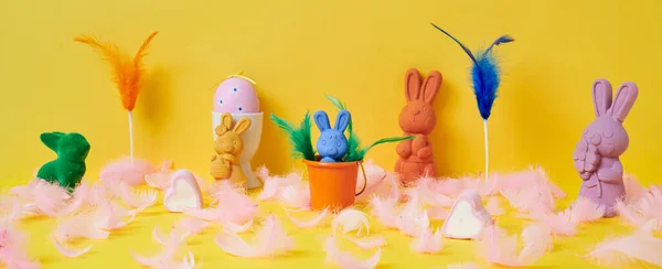 一些不同颜色的复活节兔子 一个白色蛋杯的粉色复活节蛋和一些黄色背景的粉色羽毛 以全景形式用作网页横幅或标题 — 图库照片