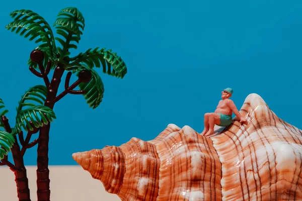 a miniature big man, wearing a green swimsuit, a green cap and a pair of sunglasses or goggles, sitting on a seashell, next to some miniature palm trees against a blue background