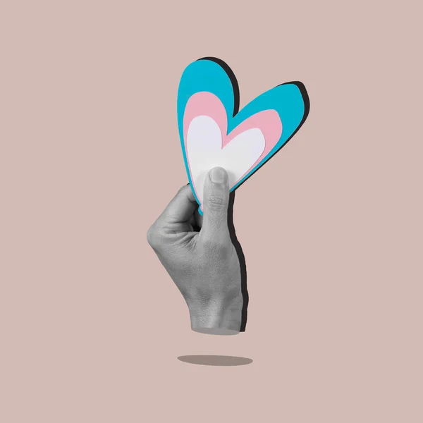 the hand of a person in black and white holding a heart with the colors of the transgender flag, on a pale brown background