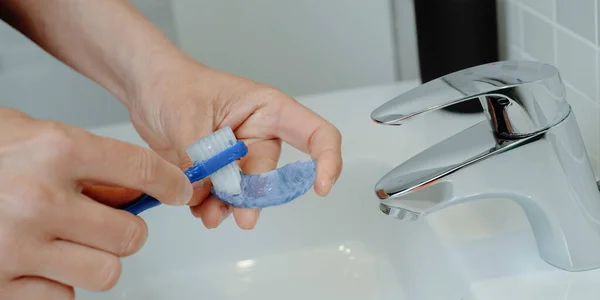 a man cleans his blue occlusal splint using a blue toothbrush in a bathroom sink, in a panoramic format to use as web banner or header