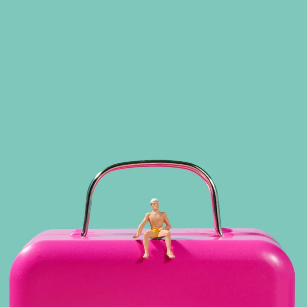 一个穿着泳衣的迷你男人坐在一个蓝色背景的粉色行李箱上 上面有一些空白的空间 — 图库照片