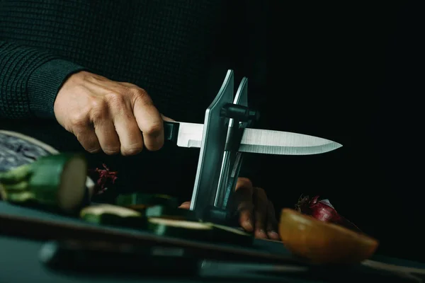 Человек Затачивает Кухонный Нож Точилкой Столе Нарезанными Нарезанными Овощами — стоковое фото