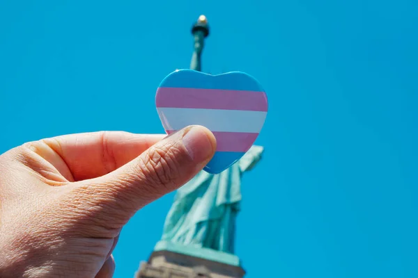 一名手持印有跨性别自豪旗的心形徽章的人在美国纽约自由女神像前的特写镜头 — 图库照片
