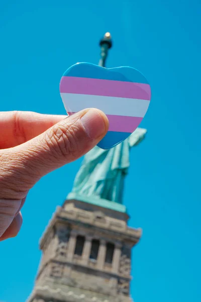 在美国纽约州自由岛的自由女神像前 一个人手持印有跨性别自豪旗的心形徽章 — 图库照片