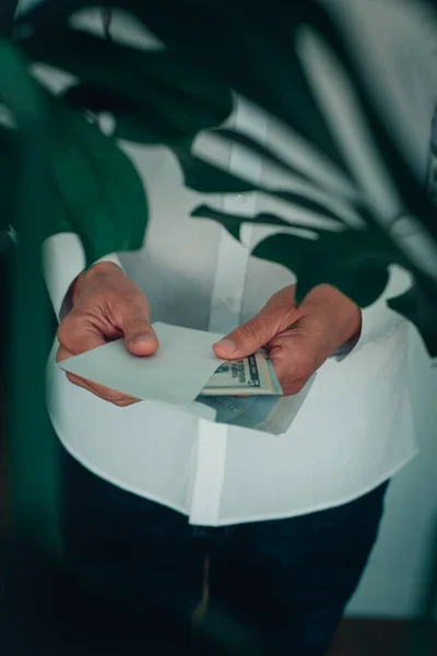 一个穿着白色长袖衬衫的男人 把一捆钞票放进或拿出了一个信封 就像从怪兽植物的叶子中看到的那样 — 图库照片