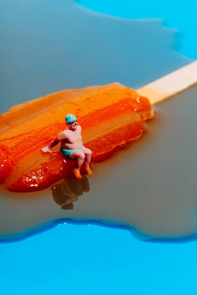 一个身披绿色泳衣 头戴泳帽 戴着一副泳镜的迷你男子的特写镜头 坐在蓝色背景的融化的橙红色冰柱上 — 图库照片