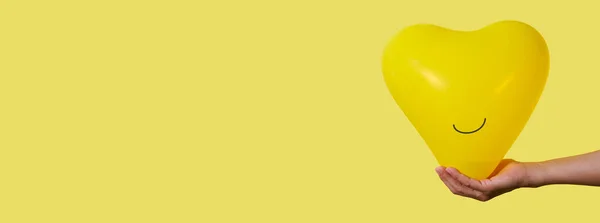 在黄色的背景上 一个男人手里拿着一个笑容可亲的黄色心形气球 左边有一些空白 是全景格式 可用作网页横幅 — 图库照片