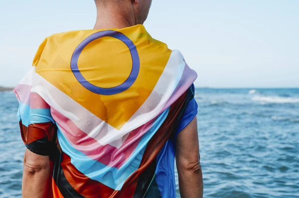 一个湿湿的人站在海里 裹着双性包容的进步的骄傲的旗帜 从后面看 — 图库照片