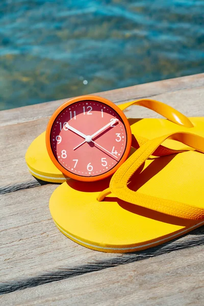 ピンクの時計と木製の桟橋の黄色のフリップフロップのペアの閉鎖は 水の横に 夏の日に — ストック写真