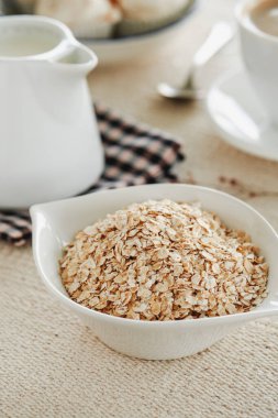 Beyaz bir seramik kasenin yanında rulo yulaf ve bej bir masa örtüsü ile birlikte sette bitki sütü bulunan beyaz seramik krema.