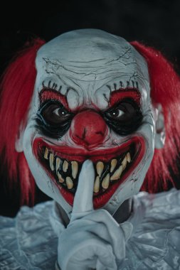 Beyaz yakalı bir kostüm giyen deli, kızıl saçlı bir palyaço, parmaklarını ağzının önüne uzatmış, sessiz olmasını dilerken, ürkütücü bir gülümseme ile gözlemciye bakıyor.