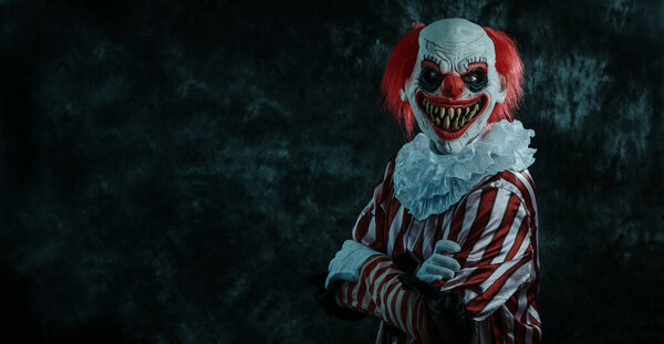 a mad evil redhead clown, wearing a white and red striped costume with a white ruff, with crossed arms and staring at the observer with a creepy smile, stands on a dark background