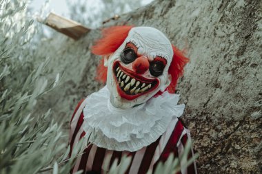 Beyaz yakalı beyaz ve kırmızı çizgili bir kostüm giyen çılgın bir kızıl saçlı palyaço, dışarıdaki eski bir evin kırsal duvarlarının önünde ürkütücü bir gülümsemeyle gözlemciye bakıyor.