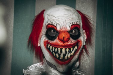 Beyaz yakalı bir kostüm giyen, sirkin yıpranmış yeşil ve beyaz çadırının önünde ürkütücü bir gülümseme ile gözlemciye bakan çılgın kızıl saçlı bir palyaçonun yakın çekimi.