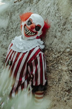 Beyaz ve kırmızı çizgili bir kostüm giymiş, eski bir evin kırsal duvarına yaslanmış çılgın bir kızıl saçlı palyaço ürkütücü bir gülümsemeyle gözlemciye bakıyor.