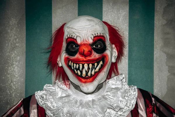 一个疯疯癫癫的红头发小丑 穿着一身白色皱领的红白相间条纹的服装 带着令人毛骨悚然的微笑凝视着马戏团的帐篷前的观赏者 — 图库照片