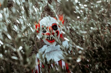 Deli bir kızıl saçlı palyaço, eski bir evin önündeki ağacın dallarının arkasında saklanan ürkütücü bir gülümseme ile gözlemciye bakıyor.