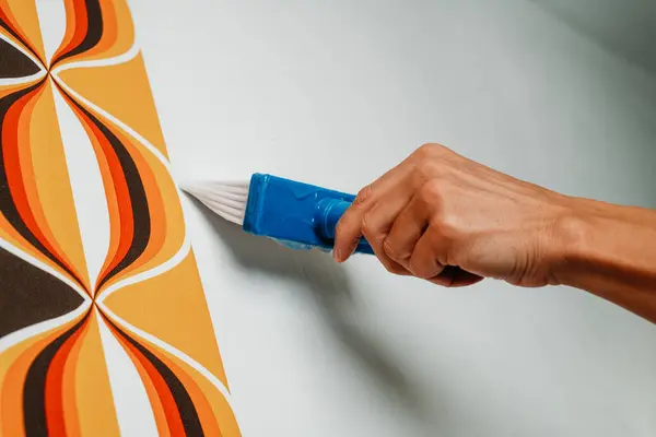 a caucasian man applies some glue with a blue brush to the wall before to attach a geometric patterned wallpaper to it