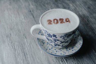 Beyaz seramik bir fincan, mavi çiçek desenli, kapuçino desenli, ve 2024 numaralı, yeni yıl sütü köpüklü, gri kırsal ahşap bir masa üzerinde